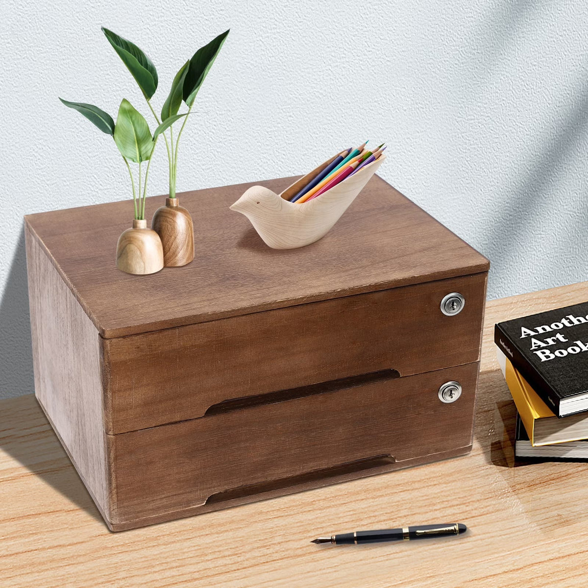 A two-drawer wooden organizer box with metal knobs sits on a tabletop. A small bird-shaped pencil holder and two mini vases with green stems rest on top.