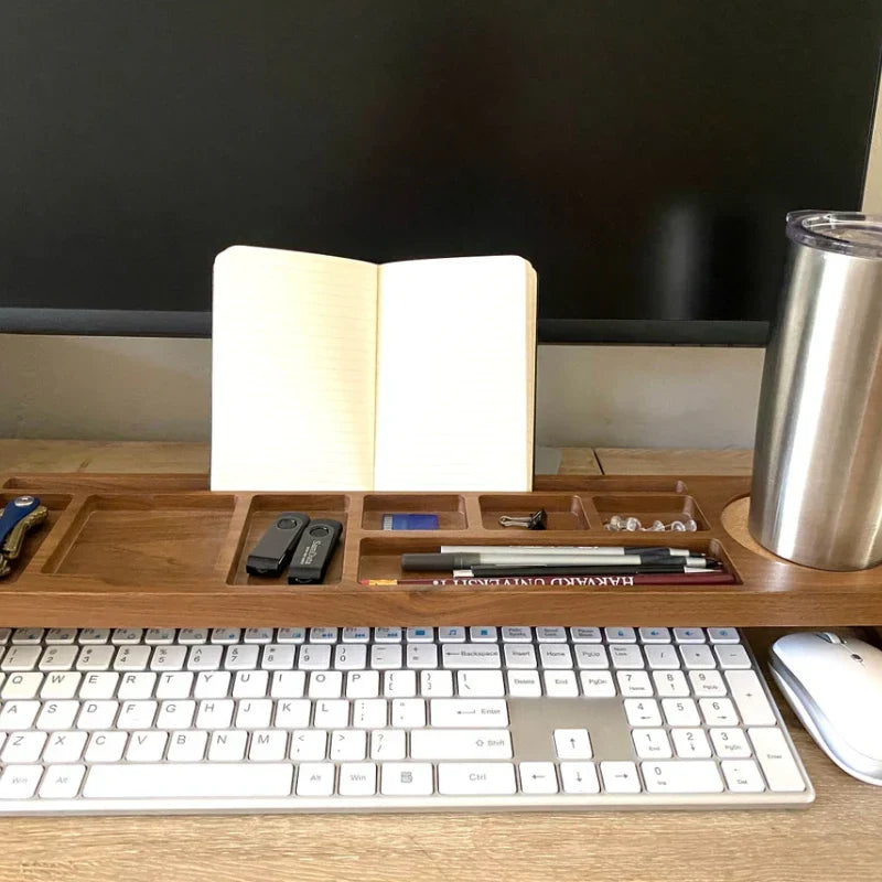 A wooden desktop organizer tray holding an open notebook, pens, a small black device, and accessories, placed on a desk in front of a keyboard and monitor with a stainless steel tumbler nearby.