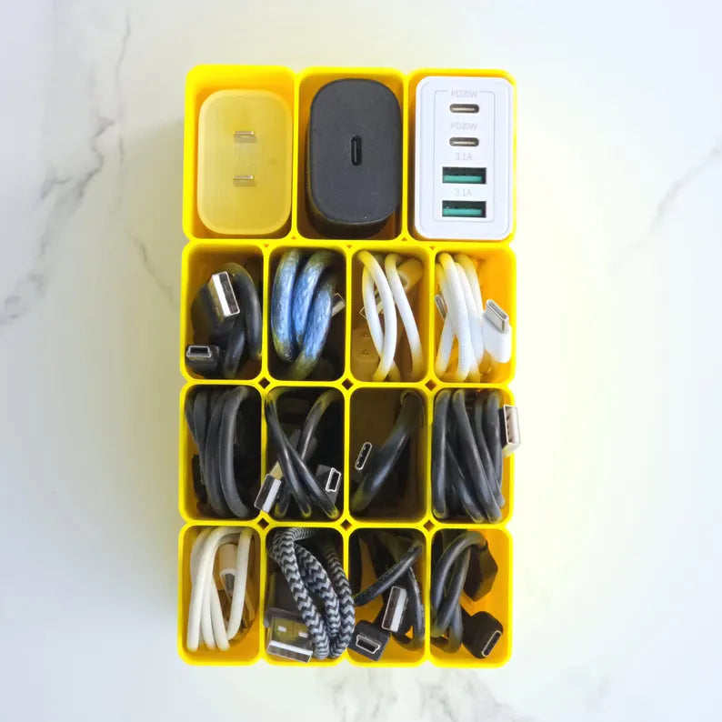 A yellow plastic organizer tray with 12 compartments, filled with various charging cables, power adapters, and USB chargers, set on a white marble surface.